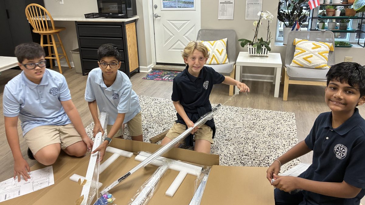 Four Middle School students kneeling on the floor around a flat-pack box, sorting parts and working through the assembly together