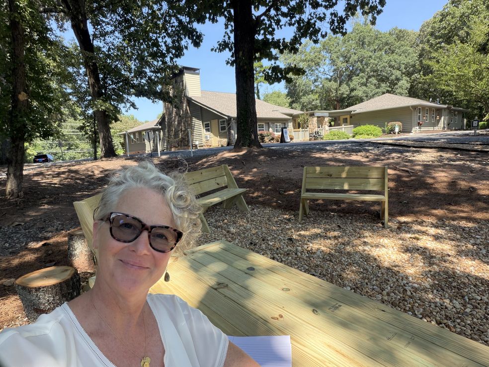Mrs. Prosser at the new picnic table in the shaded gathering area on the Upper Campus, campus buildings behind her