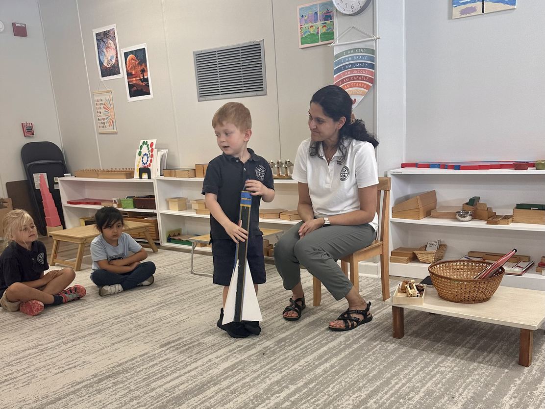 A Primary student standing before his seated classmates presenting a model rocket, his guide seated beside him