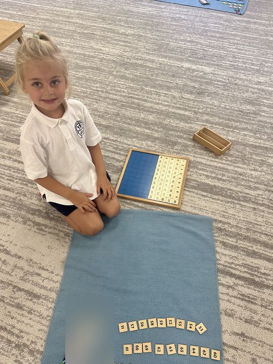 A Primary student kneeling beside the Hundred Board with numeral tiles laid out on her work rug