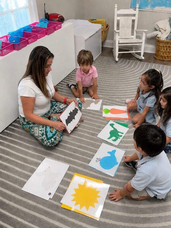 A Spanish guide seated on the rug showing silhouette cards to a circle of Primary students