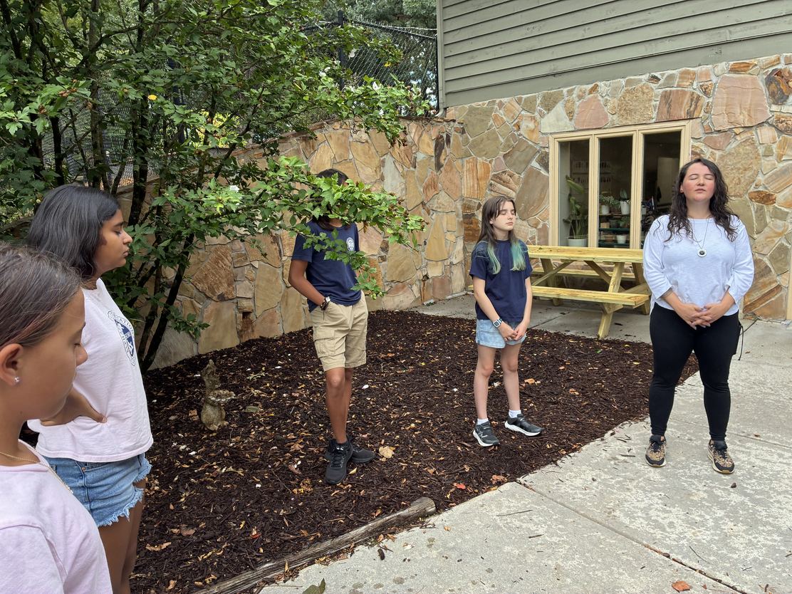 Middle School students standing outdoors with eyes closed in a moment of mindfulness beside Ms. Ramirez