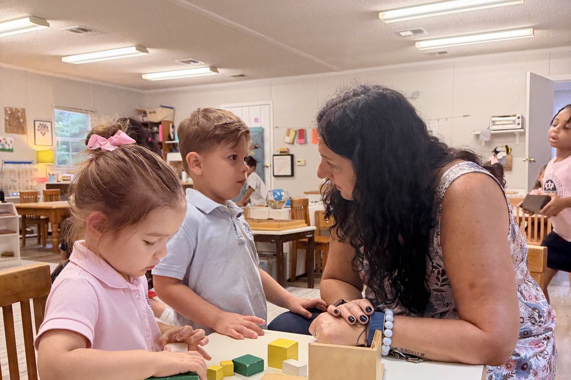 A Montessori guide giving a lesson to two children at a classroom table