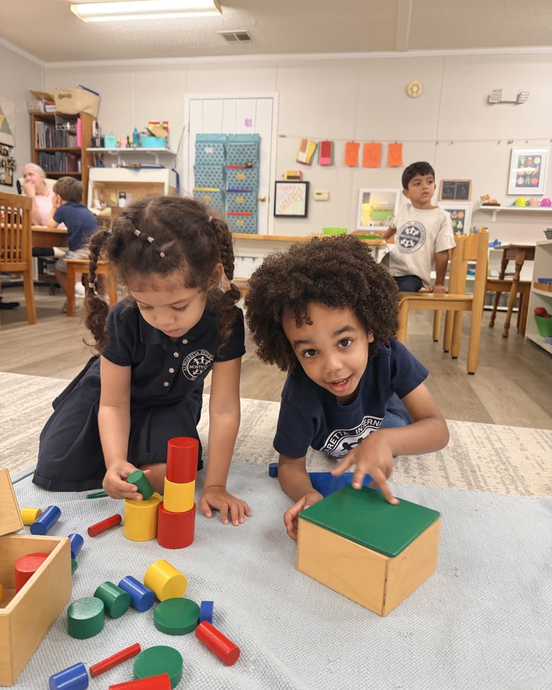 Two children exploring wooden shapes and cylinders together on a classroom mat