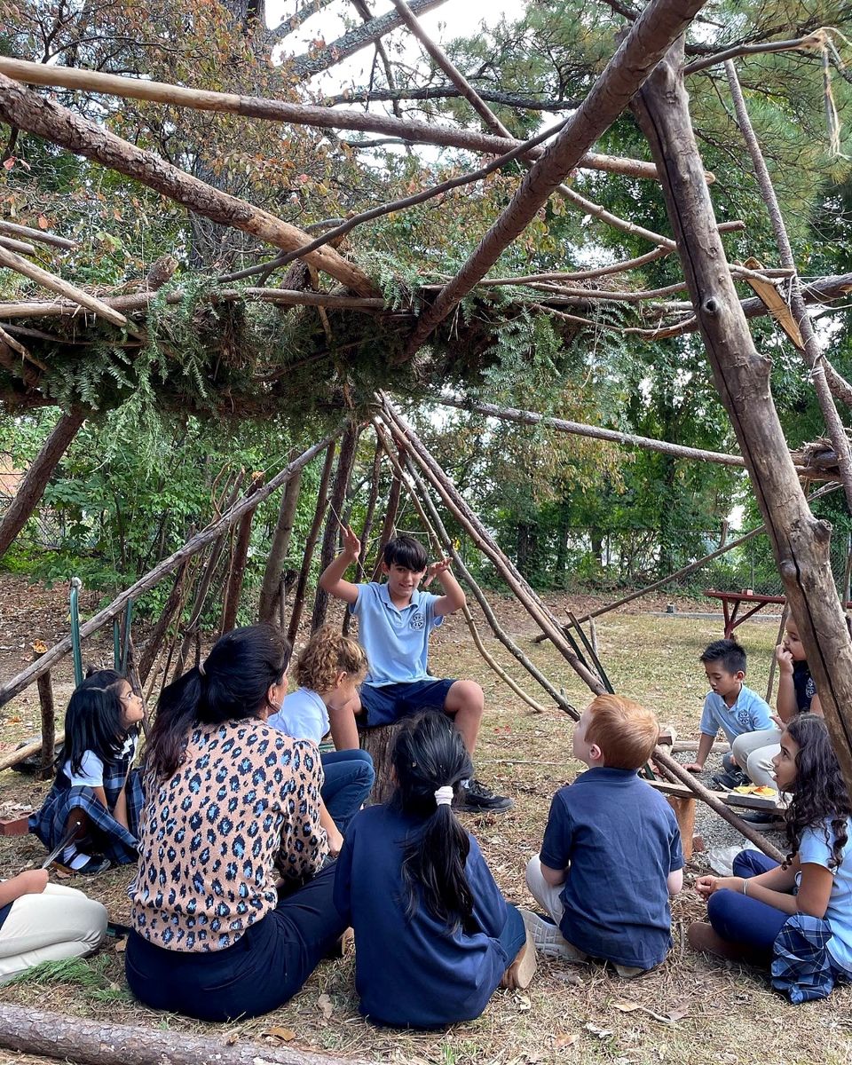 AIA students gathered with a teacher inside a stick fort they built in the wooded campus