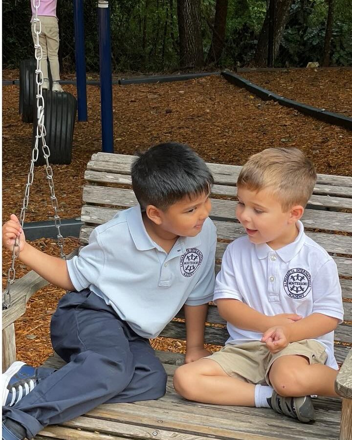 Two young AIA students in conversation on a wooden swing bench at the edge of the wooded playground