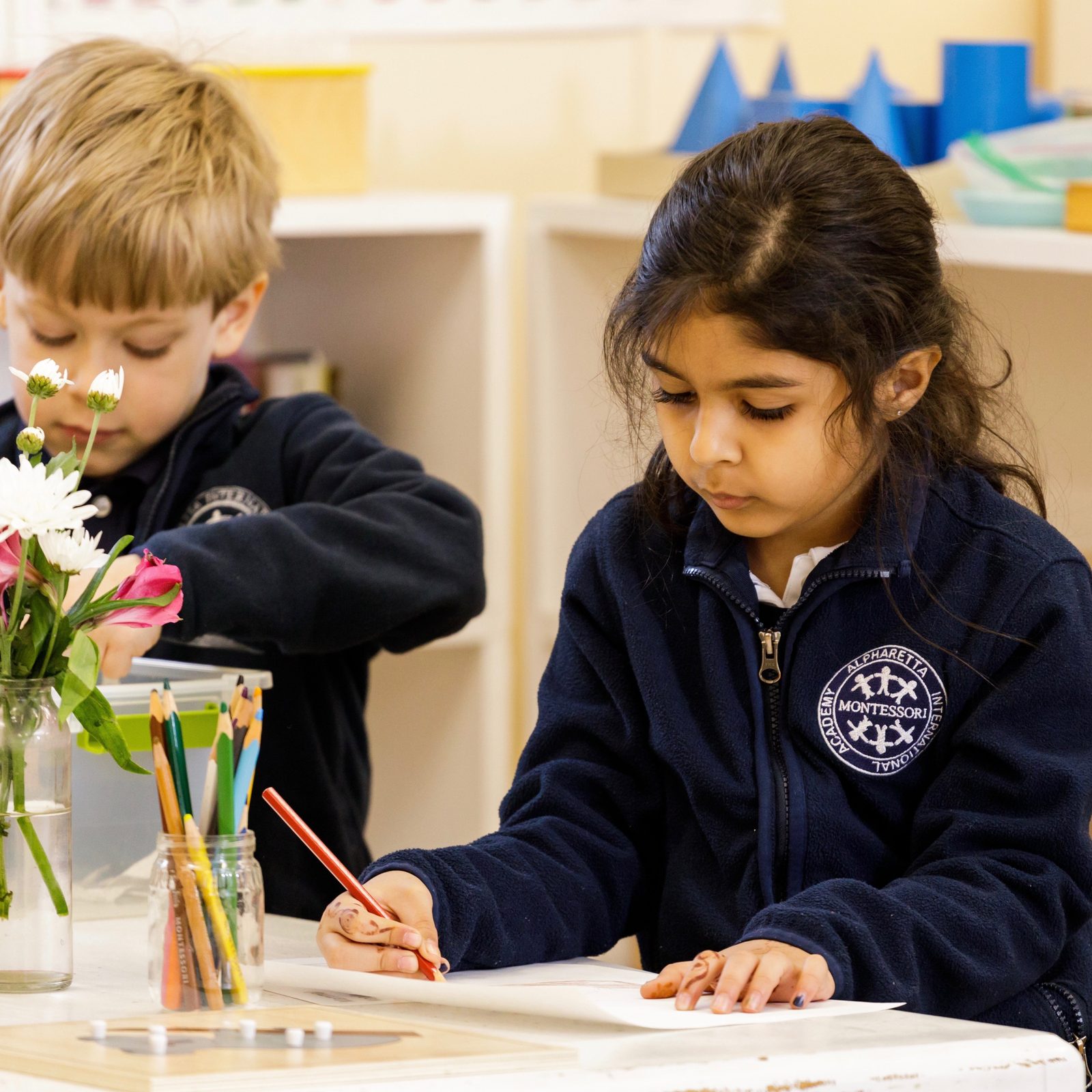 Two Primary students working side by side, one writing and one arranging flowers