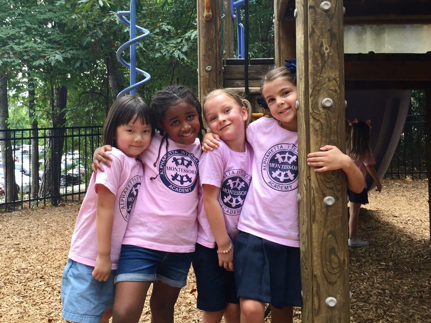 Four AIA students in matching pink school shirts on the playground, arms around each other in friendship